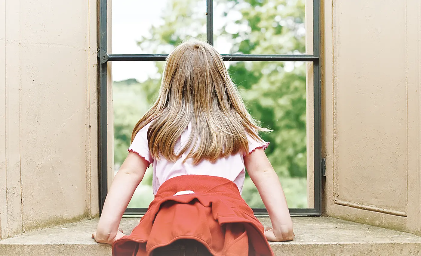 Ein Kind mit blonden langen Haaren ist von hinten zu sehen, während es durch ein großes Fenster mit Metallrahmen ins Grüne guckt.