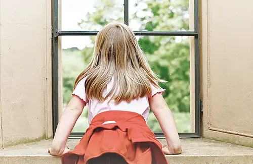 Ein Kind mit blonden langen Haaren ist von hinten zu sehen, während es durch ein großes Fenster mit Metallrahmen ins Grüne guckt.
