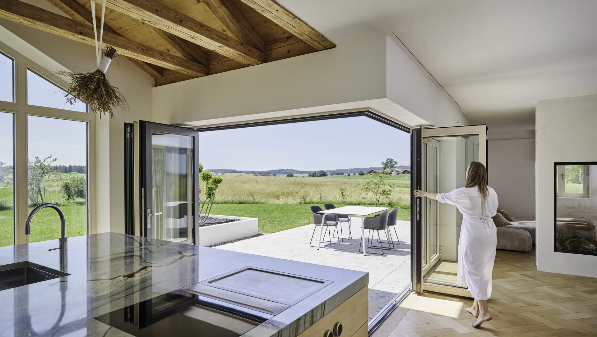 Frau öffnet Glas-Faltwand mit anthrazitfarbigem Rahmen innen und Holzrahmen innen über Eck zwischen Küche und Terrasse.