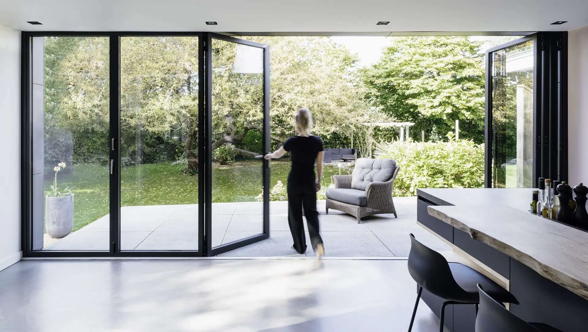 Frau öffnet Glas-Faltwand mit dunklem Rahmen zur Terrasse.