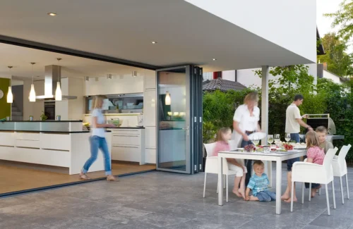 Weit geöffnete Faltanlage zwischen moderner Küche und Terasse, auf der eine Familie isst.