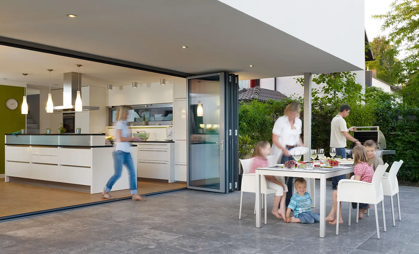 Weit geöffnete Faltanlage zwischen moderner Küche und Terasse, auf der eine Familie isst.